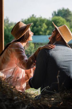 joyful woman in straw hat touching shoulder of husband near blurred hey in barn clipart