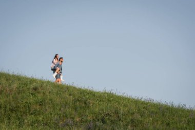 view from afar on man piggybacking girlfriend on grassy slope clipart