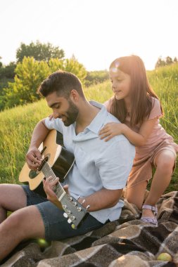 Gülümseyen bir kız babasının omuzlarına sarılıyor kırsalda dinlenirken gitar çalıyor.