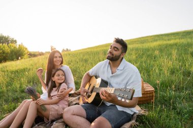 Mutlu bir kadın, kızının ve kocasının yanında gitar çalarken gülümsüyor.