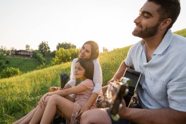 Ailesiyle kırsalda vakit geçirirken gitar çalan gülümseyen adam.