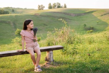 Lavanta çiçekli bir kız kırsalda bankta oturuyor ve başka tarafa bakıyor.