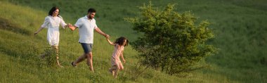 Neşeli aile el ele tutuşuyor ve yeşil yamaçlarda koşuyor.