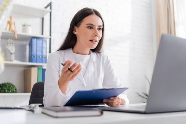 Doctor holding clipboard and talking during video call on laptop in clinic 
