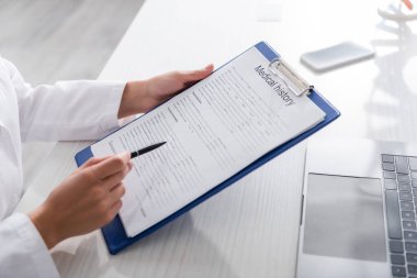 Cropped view of doctor holding medical history on clipboard near devices in clinic 