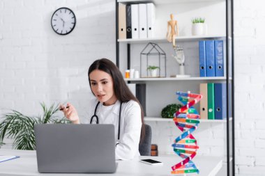 Klinikteki bulanık DNA modelinin yanında bilgisayardaki video görüşmesi sırasında kalem tutan doktor. 