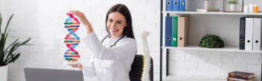 Klinikte, bilgisayarın yanında DNA modeli tutan esmer bir doktor var. 