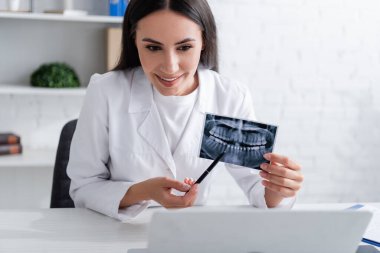 Gülümseyen doktor klinikteki dizüstü bilgisayarın yanındaki diş taramasına işaret ediyor. 