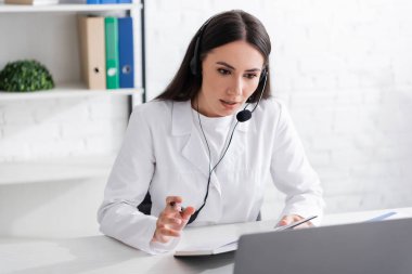 Doctor in headset holding notebook during video call on laptop in clinic 