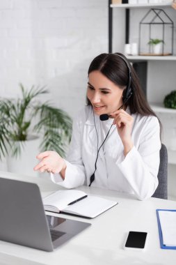 Smiling doctor in headset having video consultation on laptop near notebook and clipboard in clinic 