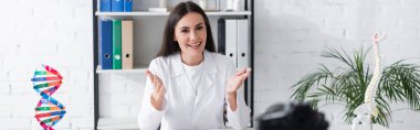 Klinikte dijital kameranın yanında konuşurken gülümseyen doktor elleriyle işaret ediyor. 