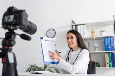 Smiling doctor pointing at clipboard near blurred digital camera in hospital 