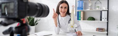 Omurilik modelinin yanında gülümseyen doktor ve klinikte bulanık dijital kamera, afiş 
