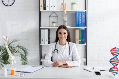 Gülümseyen doktor, klinikteki DNA ve omurilik modellerinin yakınındaki kameraya bakıyor. 