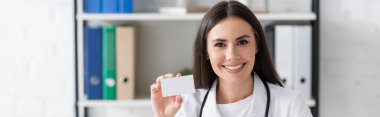 Cheerful doctor holding empty business card in hospital, banner 