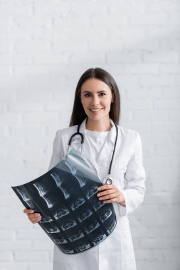Esmer doktor MRI taraması yapıyor ve klinikteki kameraya bakıyor. 
