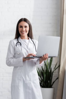Smiling doctor in white coat holding laptop and looking at camera in clinic 