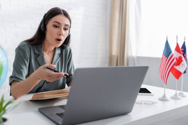 Teacher in headset talking near blurred laptop and flags on table in school 
