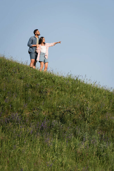 brunette woman pointing with hand while standing with man on green hill