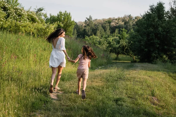 森の近くの芝生の道を走りながら手をつないでいる母親と娘の姿を — ストック写真