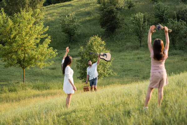 woman and girl waving hands to happy man with acoustic guitar on grassy slope