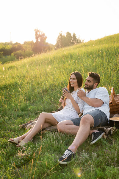 smiling man touching shoulder of woman with lavender flowers while resting on picnic in meadow