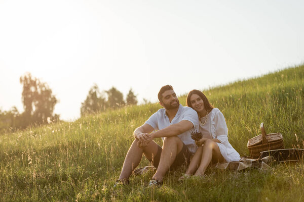 joyful couple sitting on green slope near wicker basket and looking away 