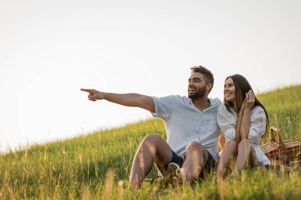 cheerful bearded man pointing away near smiling woman in green meadow 