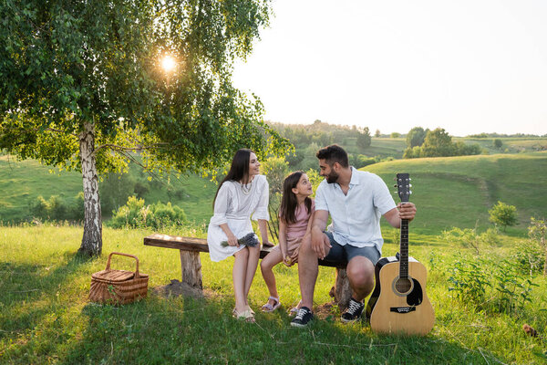 man with guitar talking to family sitting on bench under birch 