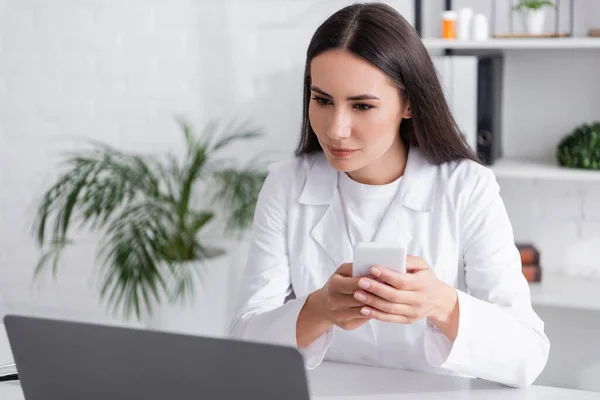Γιατρός Χρησιμοποιώντας Smartphone Και Κοιτάζοντας Laptop Στην Κλινική — Φωτογραφία Αρχείου