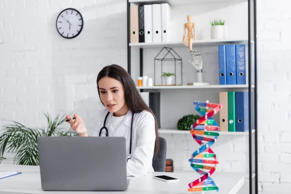 Doctor Holding Pen While Talking Video Call Laptop Blurred Dna — Stock fotografie