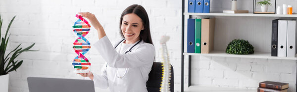 Positive brunette doctor holding dna model near laptop in clinic, banner 