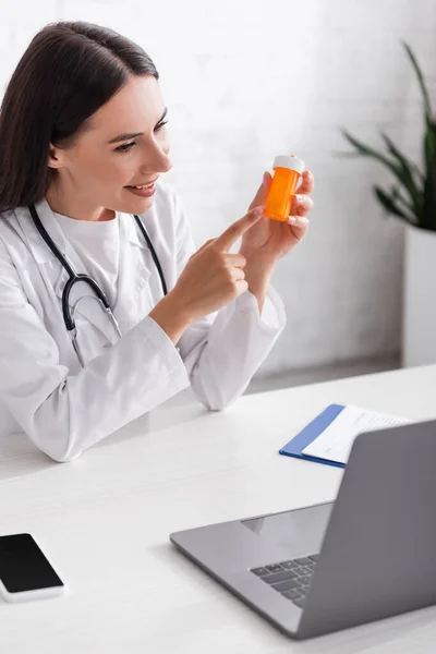 Médico Positivo Apontando Para Frasco Com Pílulas Durante Videochamada Laptop — Fotografia de Stock