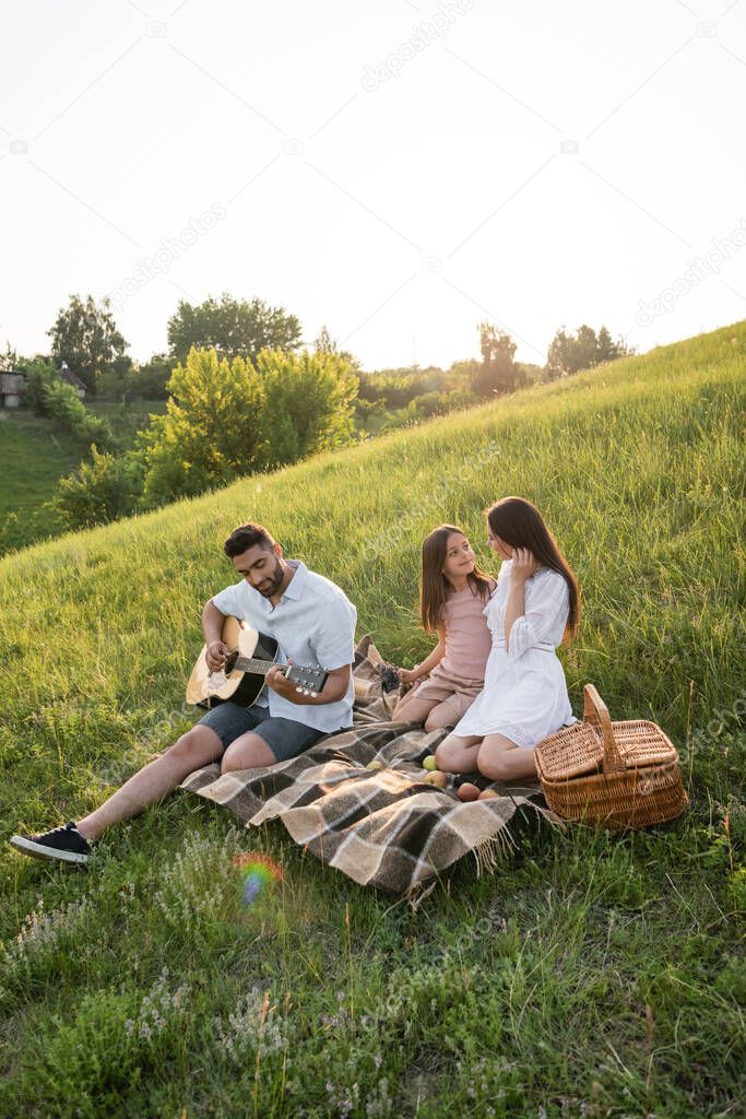 man playing guitar near family sitting on plaid blanket in green meadow