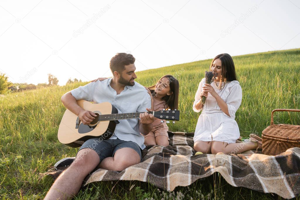 woman smelling aromatic lavender near daughter and husband playing guitar in green meadow
