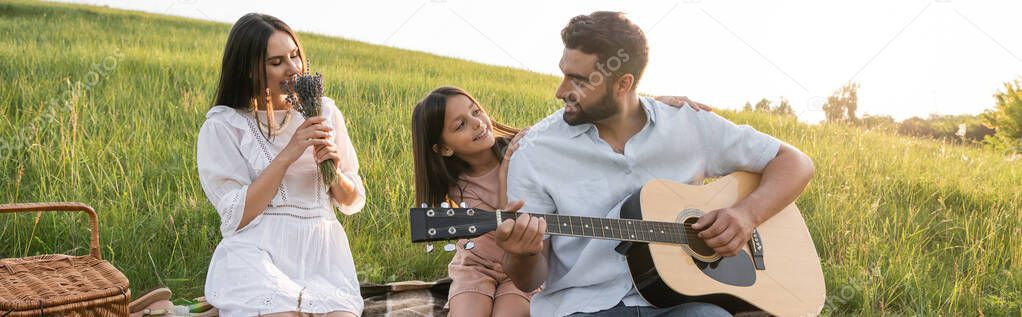 pleased woman smelling lavender bouquet near daughter and husband playing guitar, banner