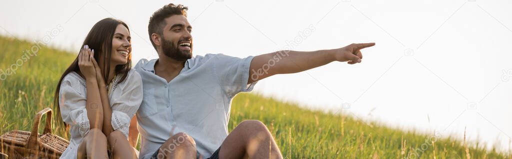 cheerful man pointing with finger near woman while sitting on slope in field, banner