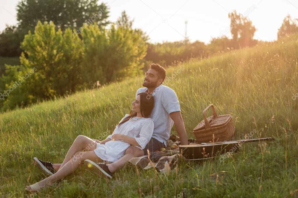 cheerful couple looking away while sitting on green slope near wicker basket and acoustic guitar