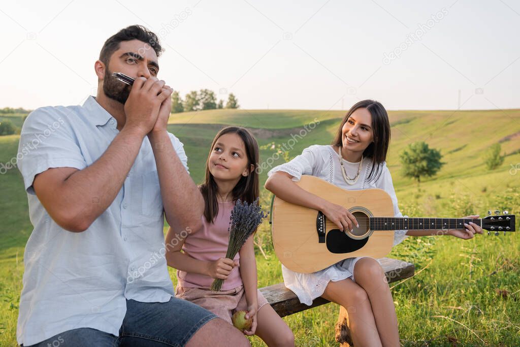 man playing harmonica near wife with guitar and happy child in countryside