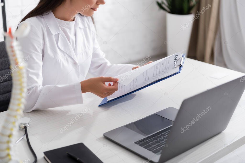 Cropped view of doctor holding clipboard near laptop and stethoscope in clinic 