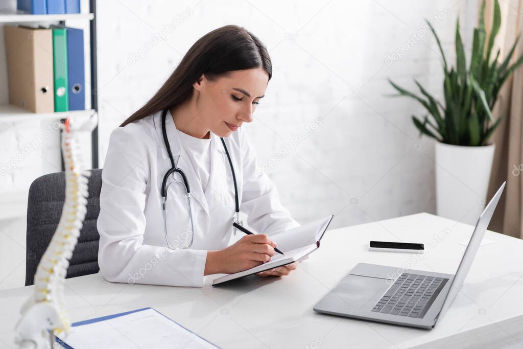 Doctor writing on notebook near devices and blurred spinal model in clinic 