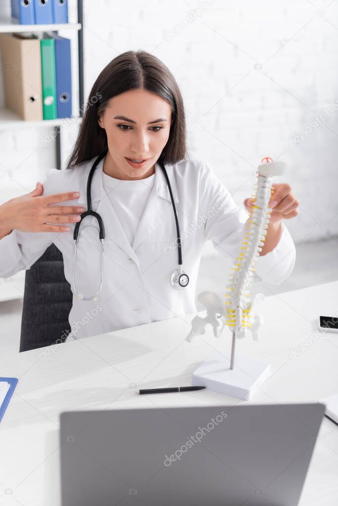 Brunette doctor holding spinal model during online consultation on laptop in hospital 