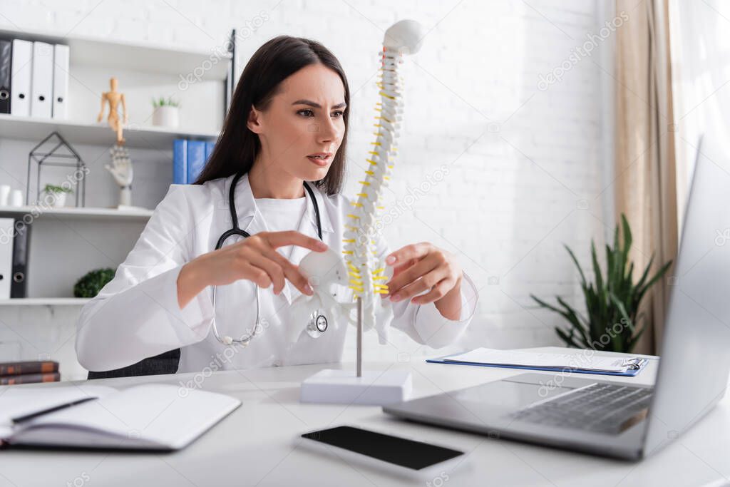 Doctor holding spinal model during online consultation on laptop in clinic 