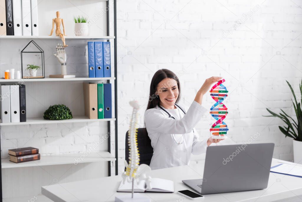 Cheerful doctor in white coat holding dna model during online consultation on laptop in clinic 