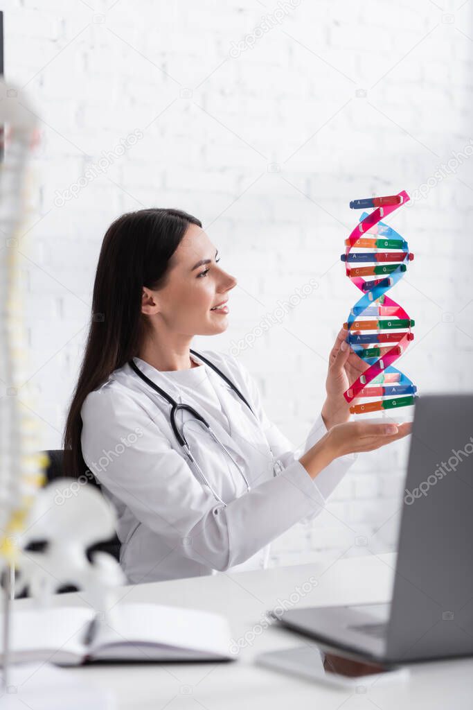 Side view of cheerful doctor holding dna model near blurred gadgets and notebook in clinic 