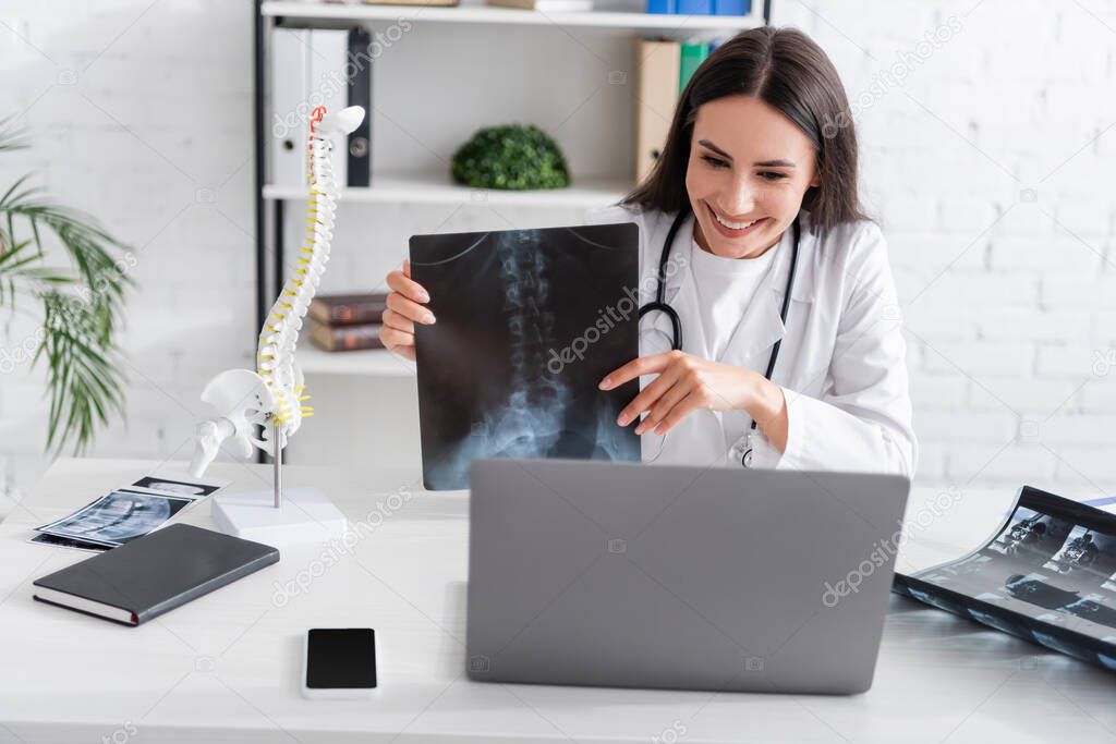 Positive doctor holding x-ray scan during online consultation on laptop in hospital 