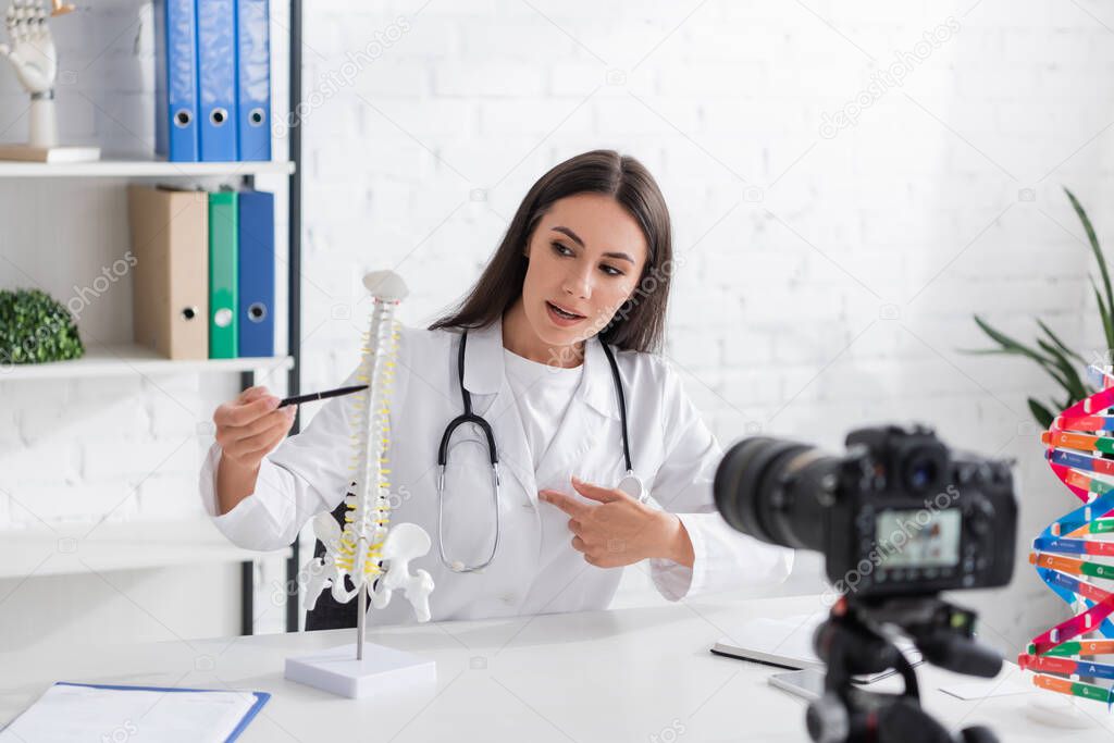 Brunette doctor pointing at spinal model near blurred digital camera in clinic 