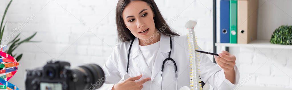 Doctor in white coat pointing at spinal model near digital camera in clinic, banner 