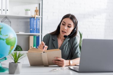 Teacher in headset holding book during online lesson on laptop in school 