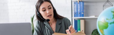 Brunette teacher in headset reading book near laptop and blurred globe in class, banner 